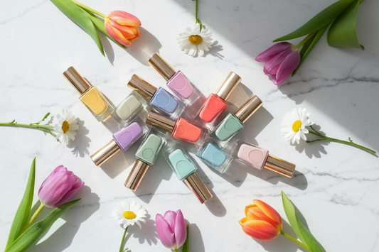 "Spring 2026 trending nail polish colors including butter yellow, lavender, coral, sage green, sky blue, peach, lilac, mint, orange, and blush pink arranged on white marble with fresh spring flowers